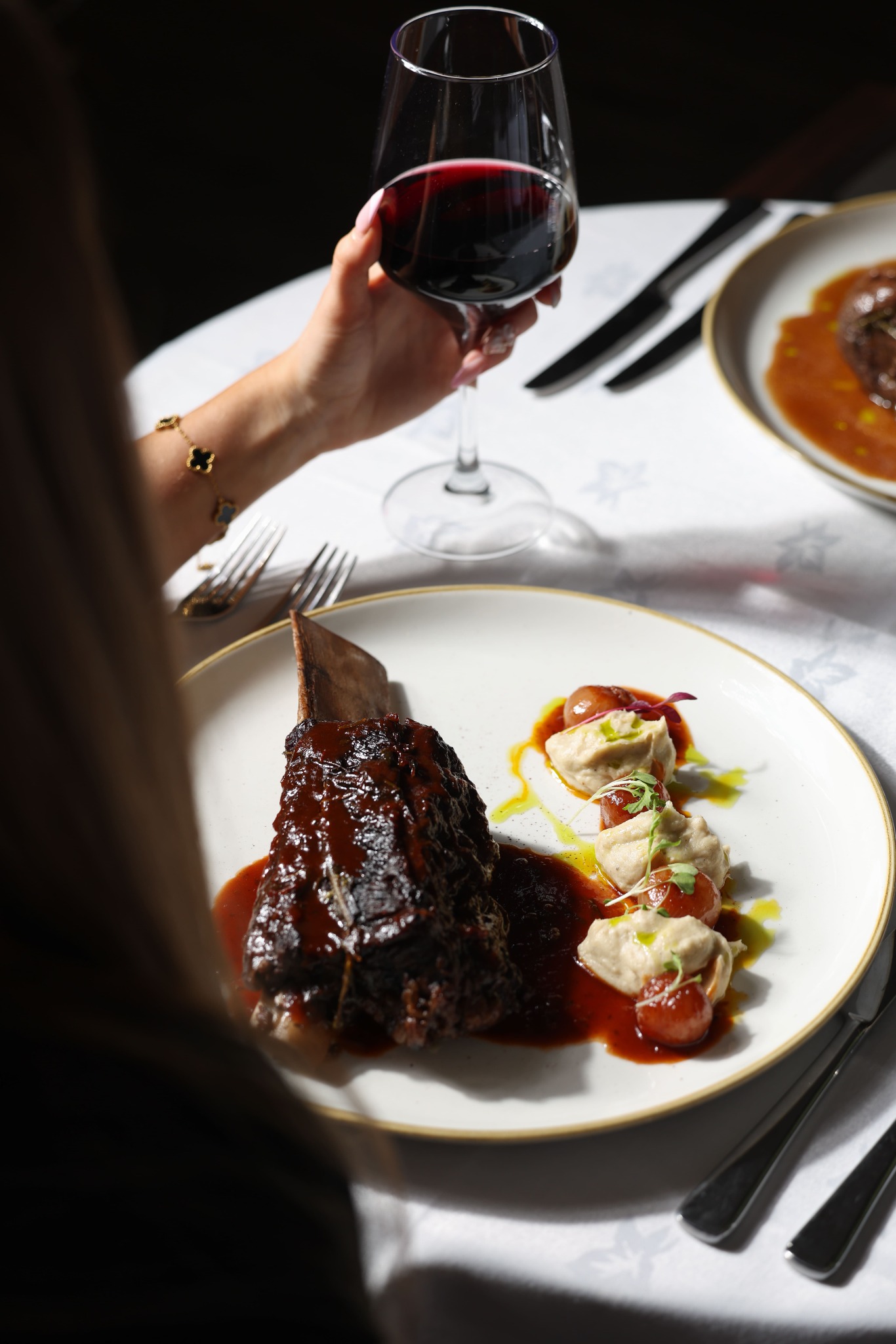 A guest with a glass of red wine beside a richly glazed main dish at Yammas
