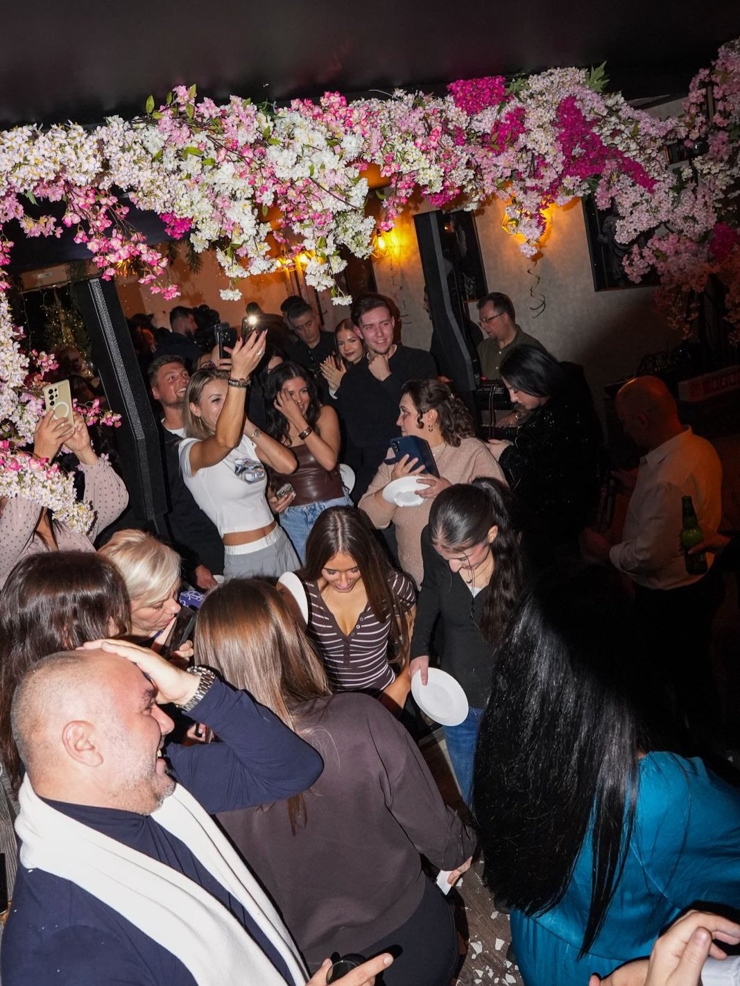 Guests enjoying a lively Greek night at Yammas Greek Restaurant in Beckenham, dancing under floral decorations with plate smashing and a vibrant party atmosphere.